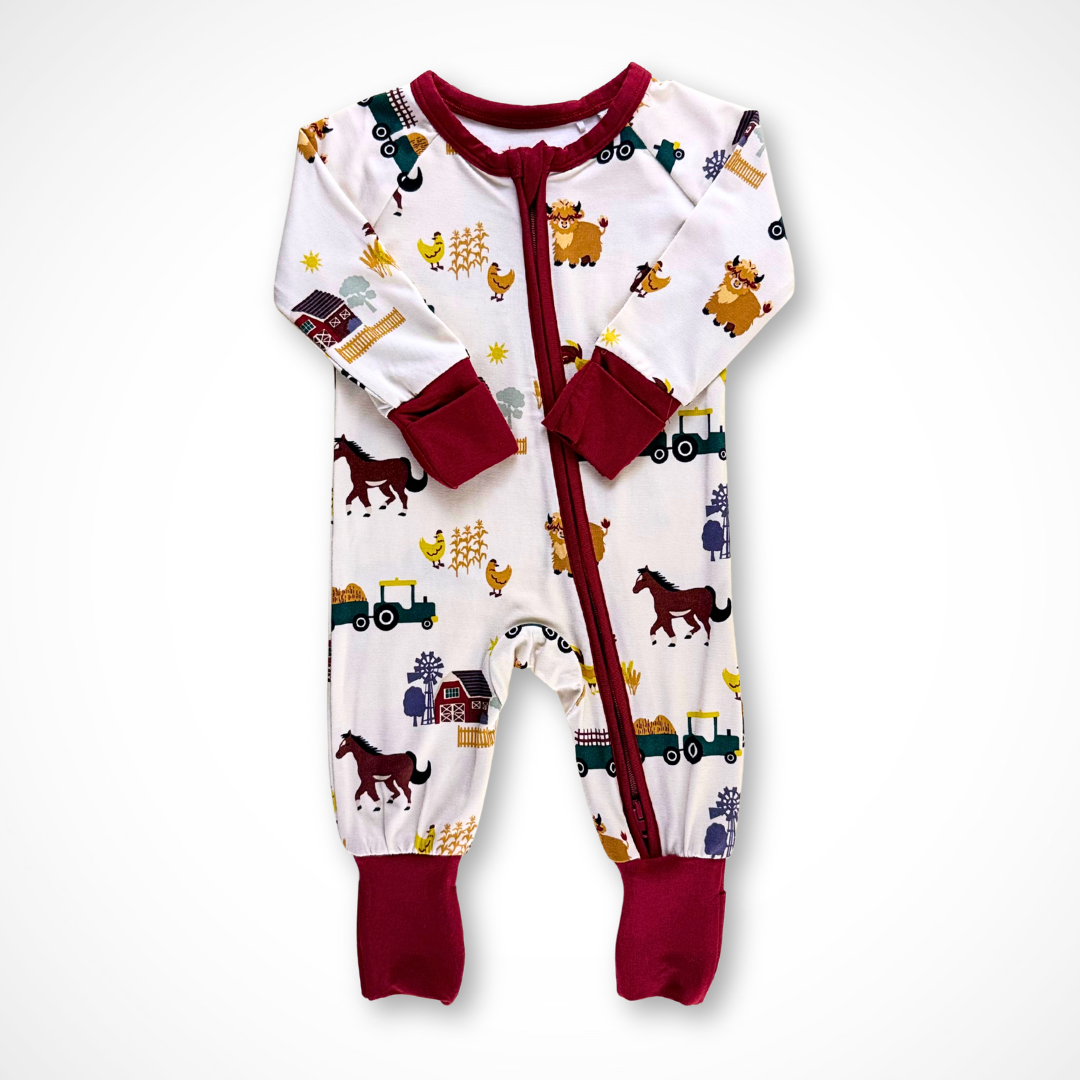 Long-sleeve baby footie pajama made from bamboo fabric. The On the Farm print features tractors, horses, highland cows, chickens, barns, and haystacks on a light taupe background, with burgundy trim and cuffs. 