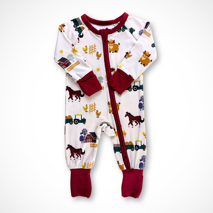 Long-sleeve baby footie pajama made from bamboo fabric. The On the Farm print features tractors, horses, highland cows, chickens, barns, and haystacks on a light taupe background, with burgundy trim and cuffs. 